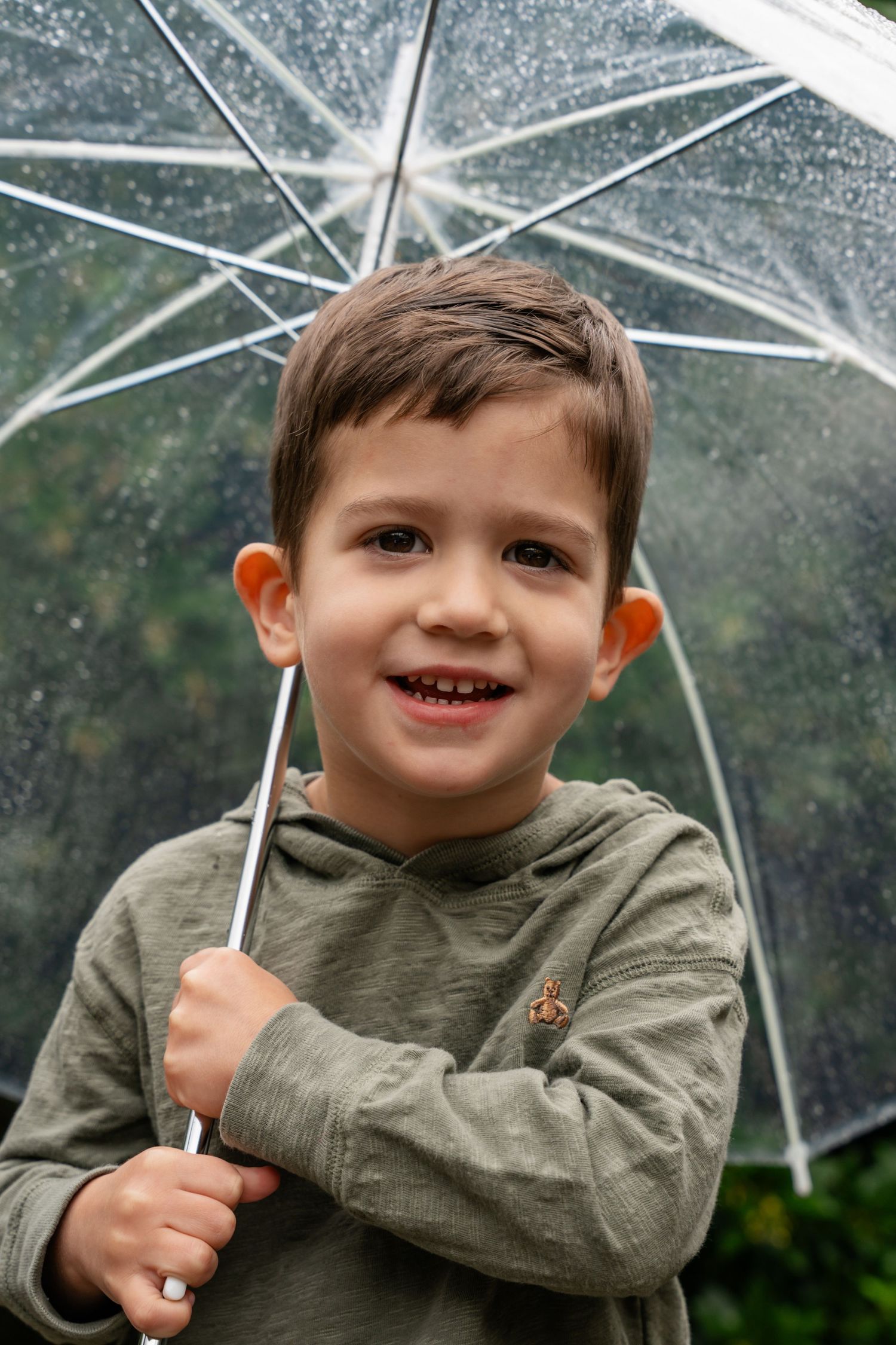 Happy young boy holding a clear umbrella in the rain, wearing a green hoodie, smiling at camera.