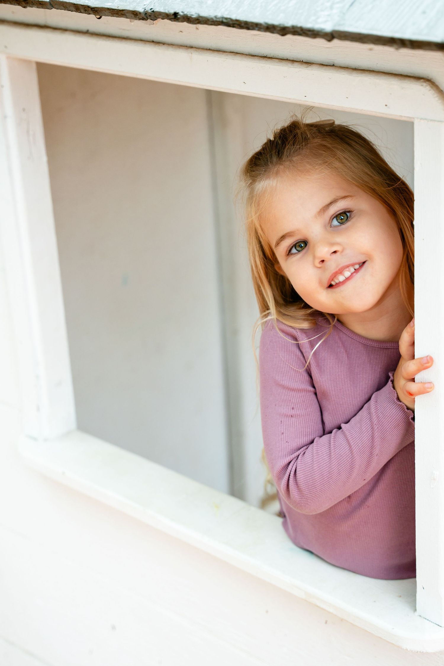 Young smiling girl with blonde hair wearing a purple top, peeking through a white window frame.