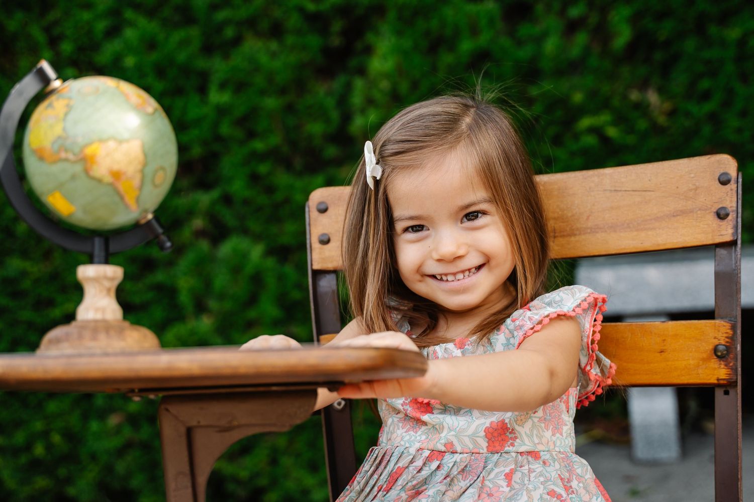 Smiling toddler girl in floral dress sitting at wooden desk outdoors with globe.