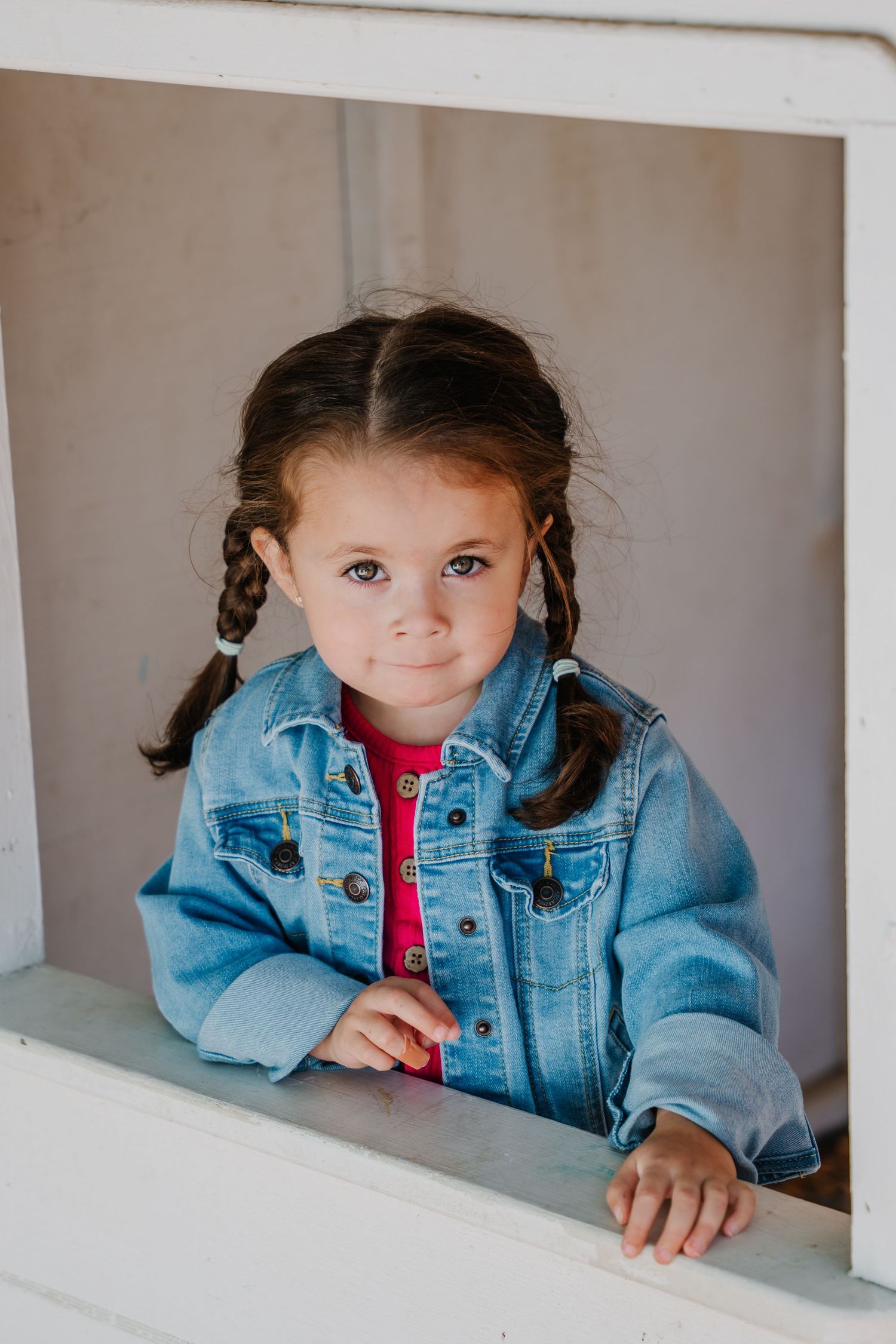 Young girl with pigtails wearing a denim jacket and pink shirt, leaning on a white window ledge.