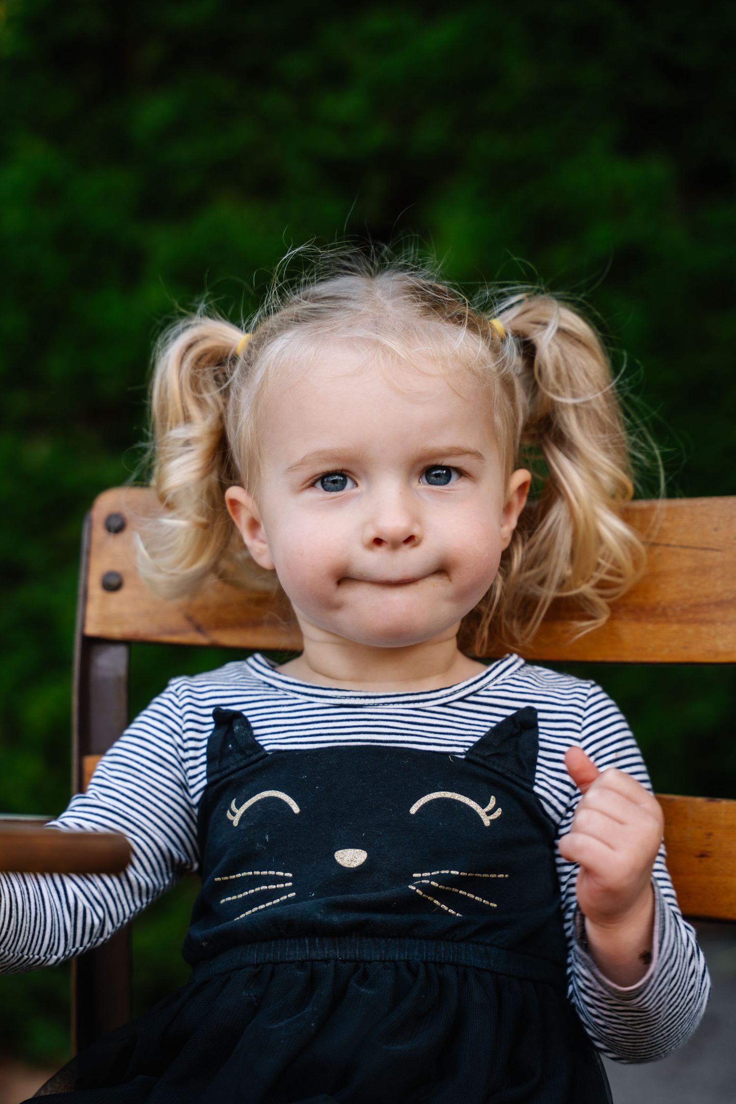 Cute blonde toddler girl with pigtails wearing a black cat dress sits on a wooden chair outdoors.