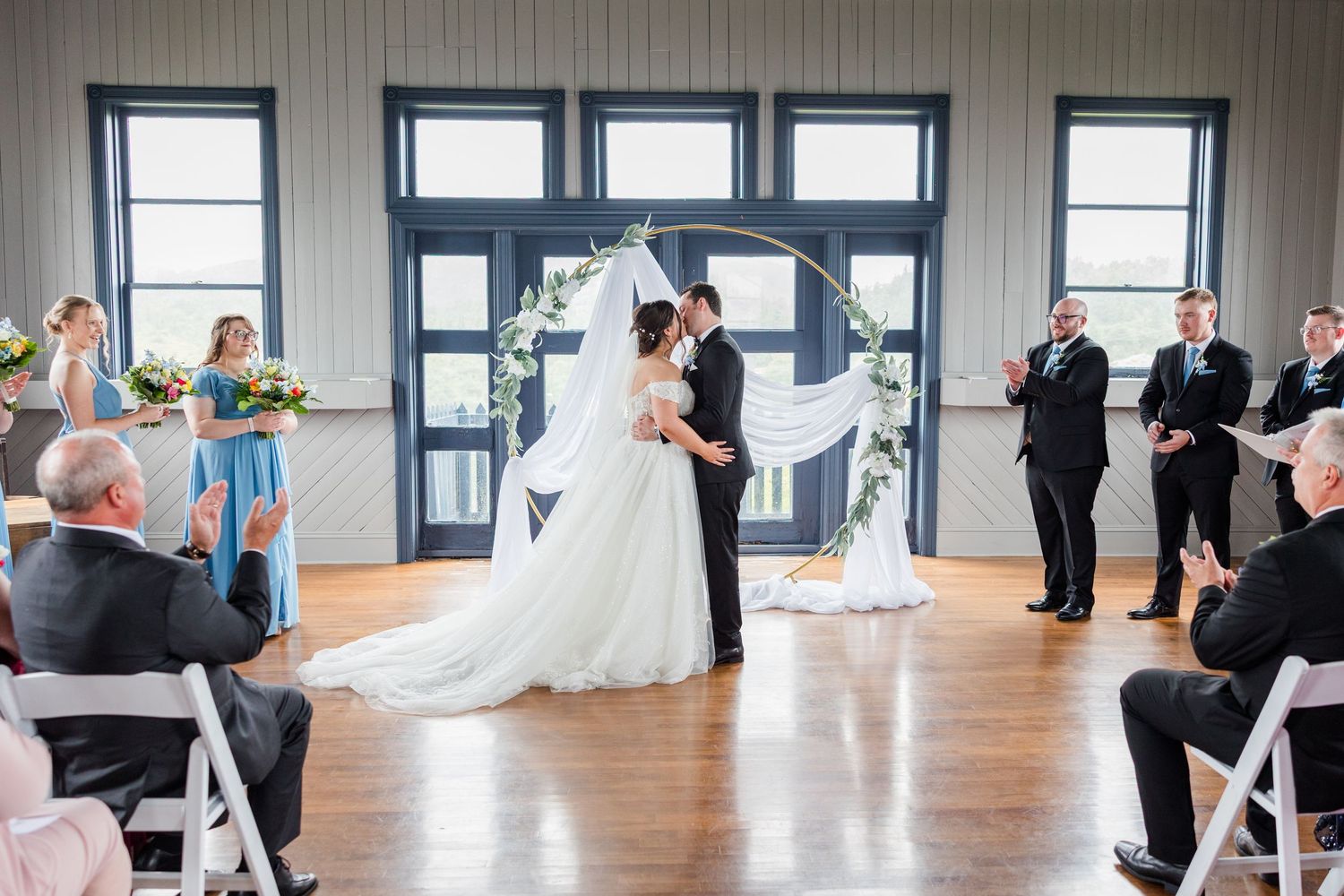 Bride and groom exchange vows at elegant indoor wedding ceremony with circular floral arch and white chairs.