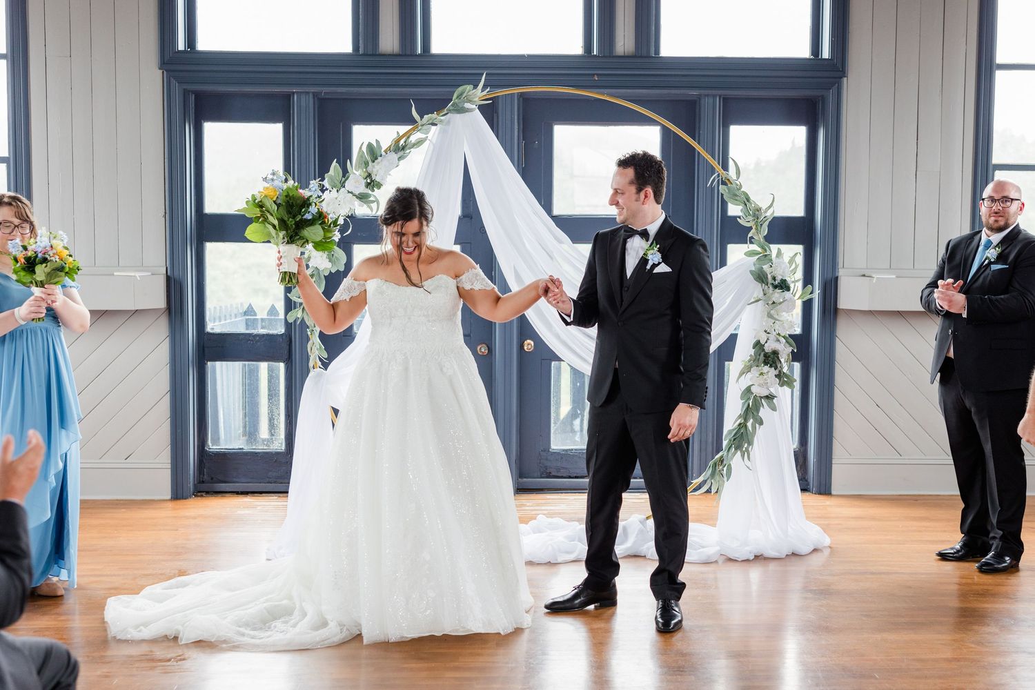 Bride and groom celebrate under a circular floral arch during their indoor wedding ceremony with blue-dressed bridesmaids.