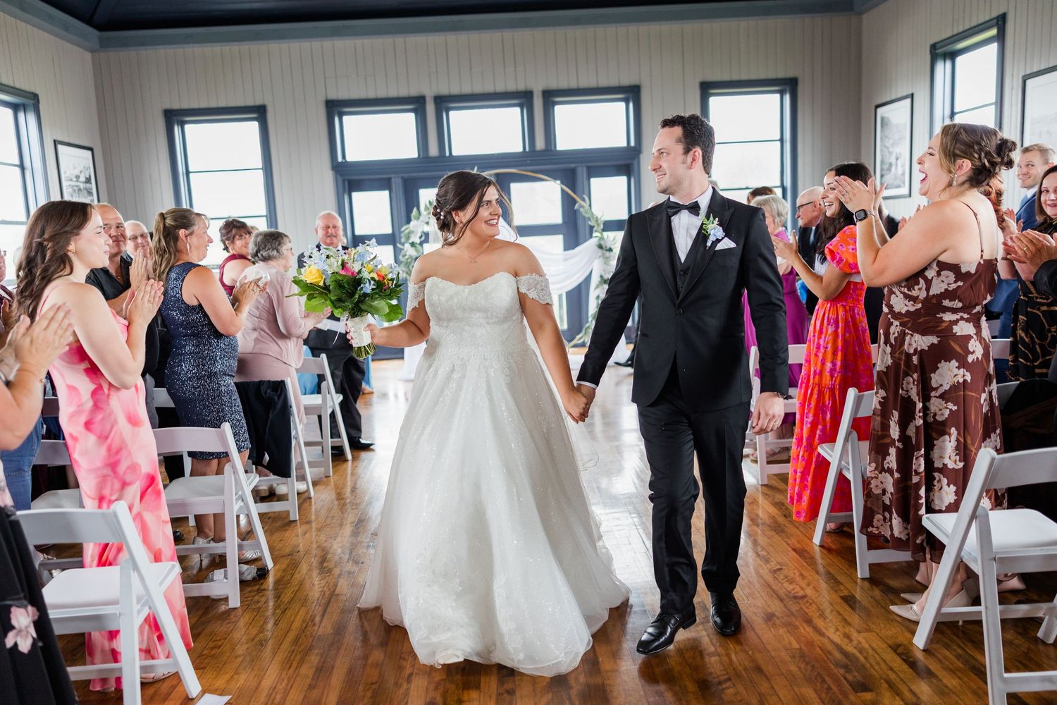 Bride and groom walk down the aisle at indoor wedding ceremony as guests celebrate with applause.