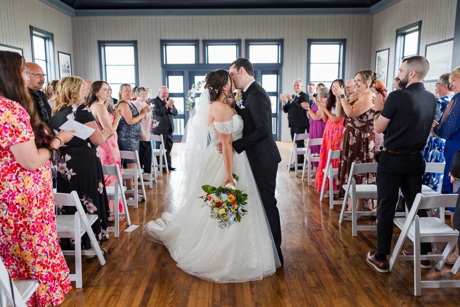 Bride and groom share first kiss at wedding ceremony while guests applaud in elegant white chair-lined aisle.