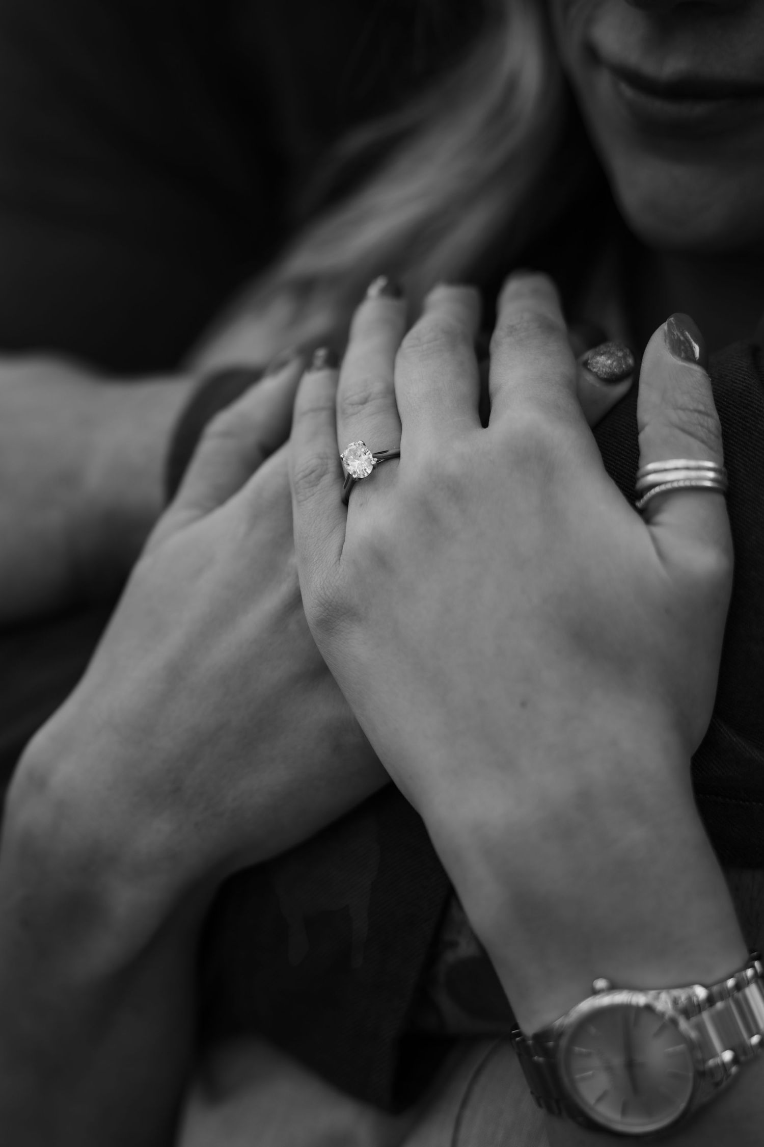 Black and white photo of couple's hands showing engagement ring, bracelet, and watch in romantic embrace.
