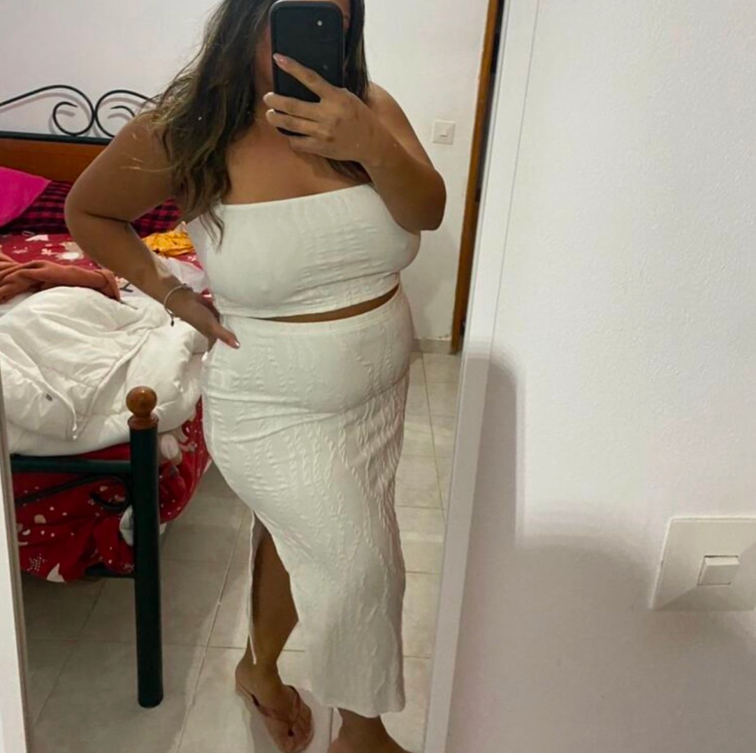 Woman in white crop top and midi skirt set posing in mirror selfie with phone.