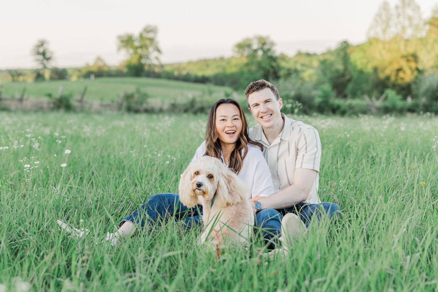 Happy couple sitting in a lush green field with their fluffy golden dog during a sunny outdoor engagement photo session.