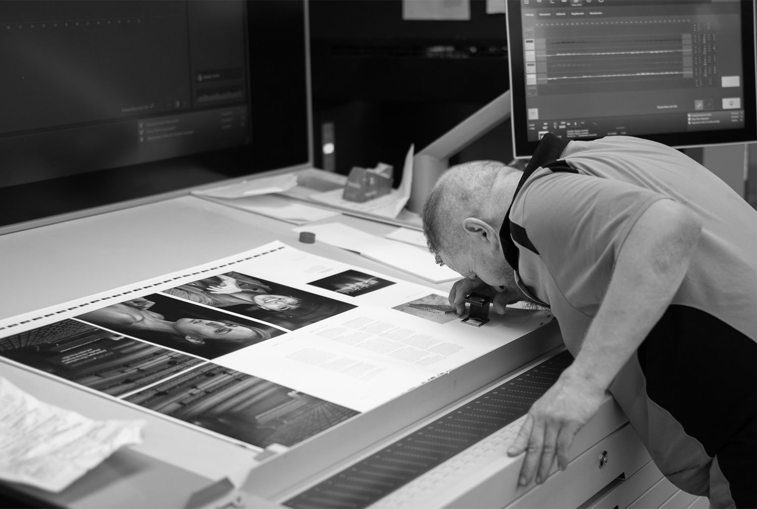 Black and white photo of a man closely examining large printed photographs on a light table near computer monitors.