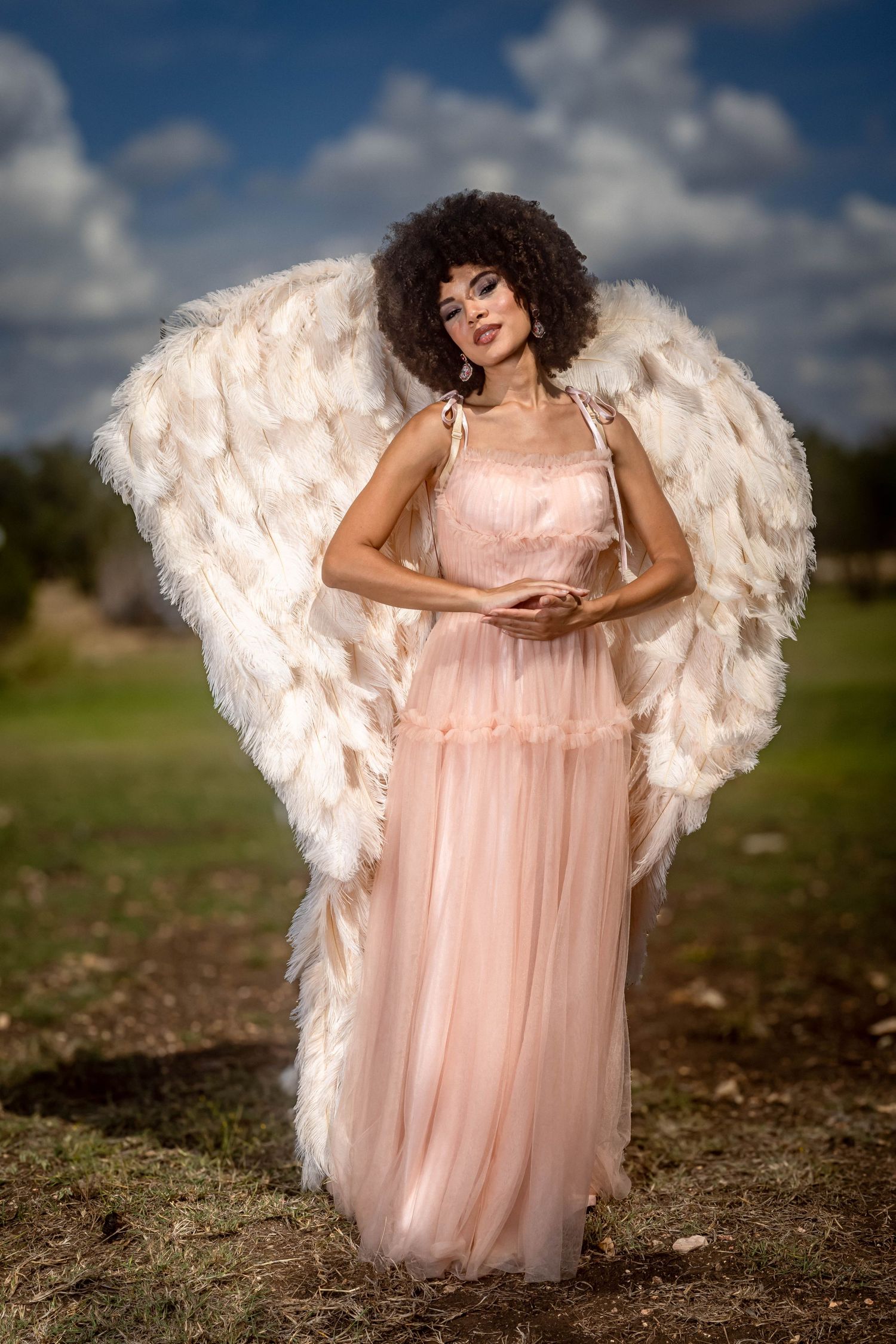 Woman in pink gown with large white feather angel wings poses outdoors under cloudy blue sky.