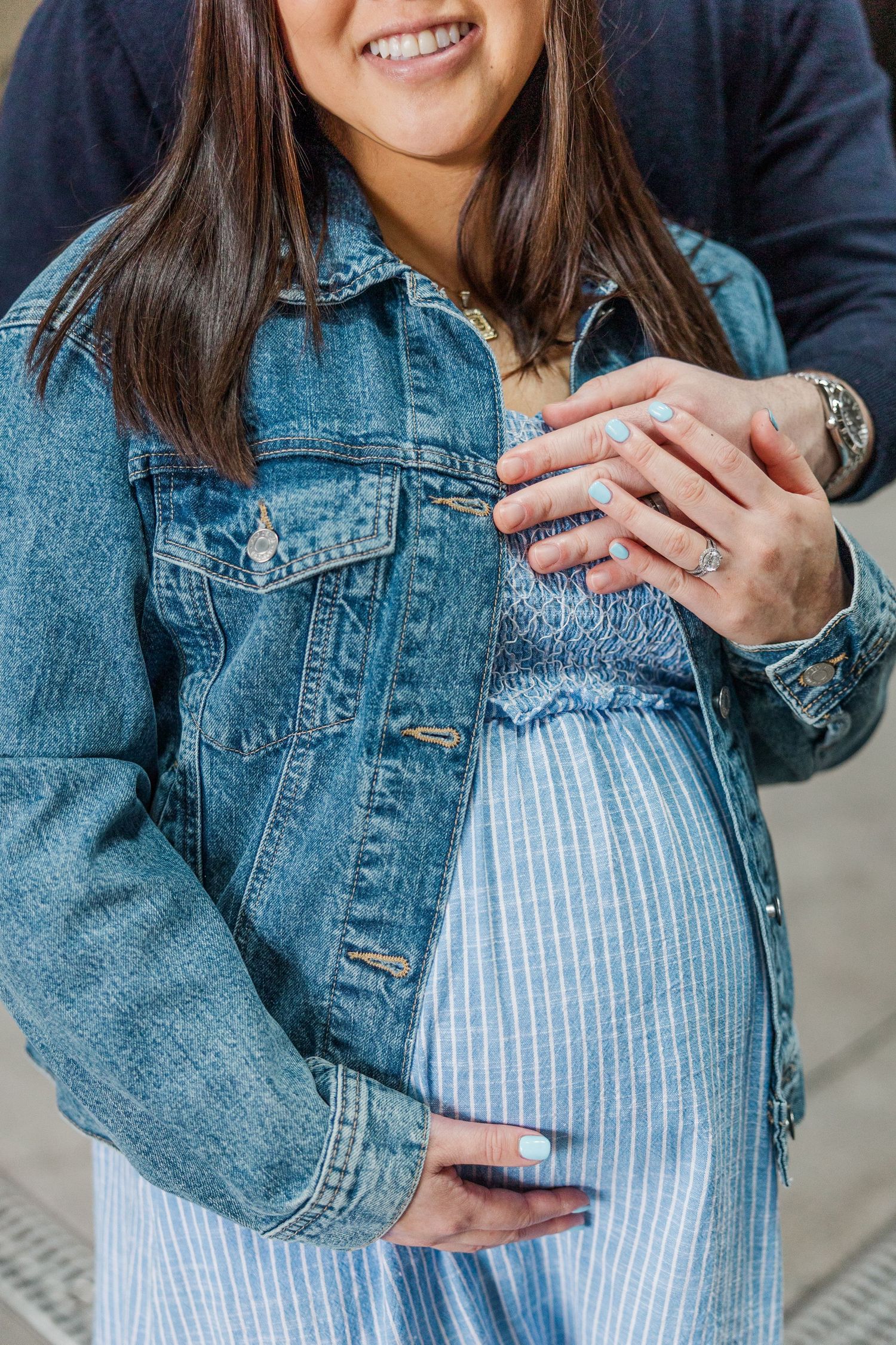 Pregnant woman in denim jacket and striped dress holding baby bump, smiling during maternity photo session.