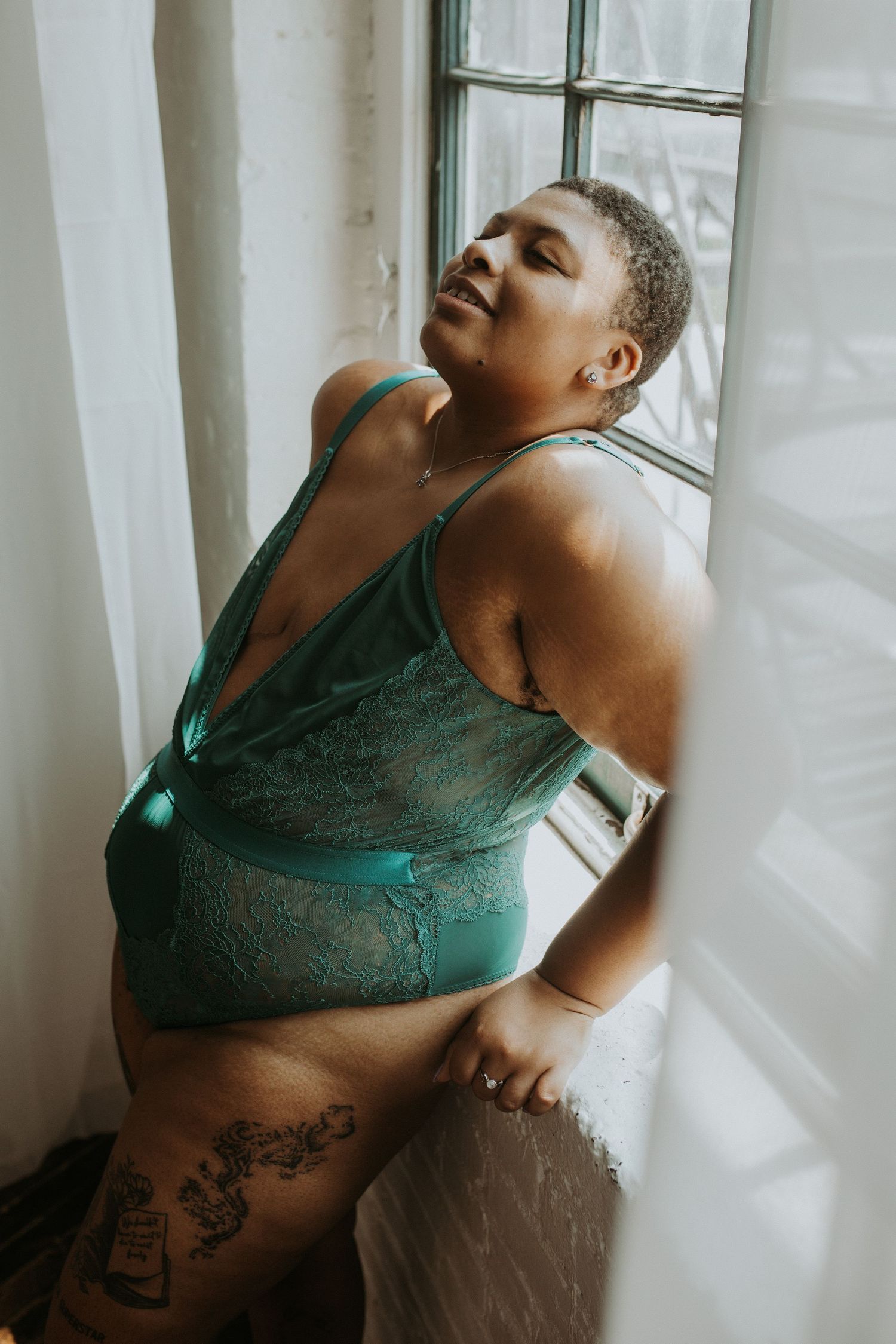 Plus-size woman with tattoos smiling confidently in black lace lingerie, sitting on a bed in a bright loft.