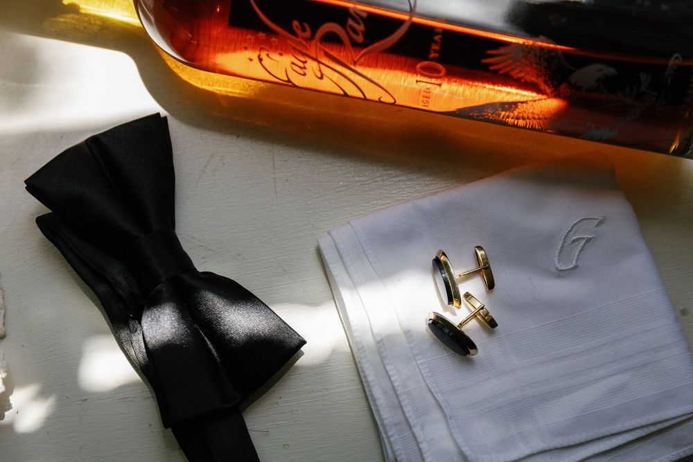 Black bow tie, gold cufflinks, monogrammed white handkerchief, and amber bottle arranged on white surface.