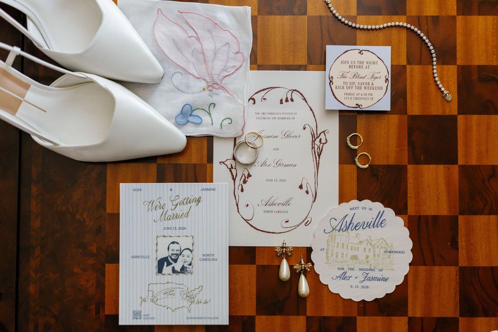 Flat lay of wedding stationery suite with invitations, rings, earrings, and white bridal heels on wood floor.