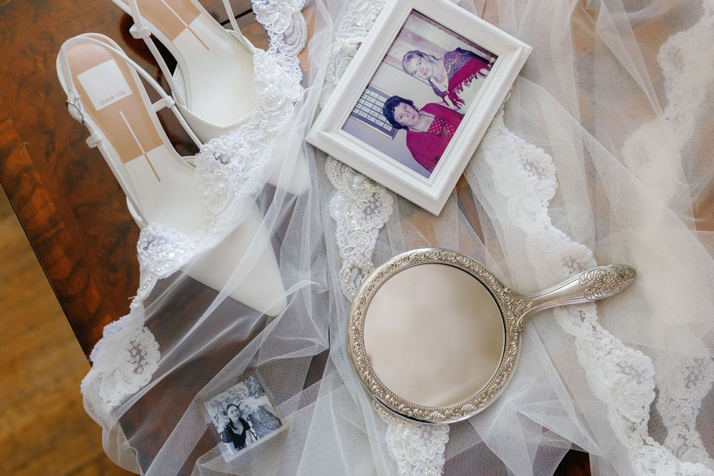Bridal flat lay with lace veil, white heels, hand mirror, and framed family photo on wooden surface.