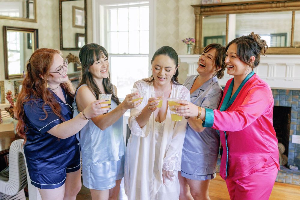 Bride in white robe toasting champagne with four bridesmaids in colorful pajamas during wedding morning preparations.
