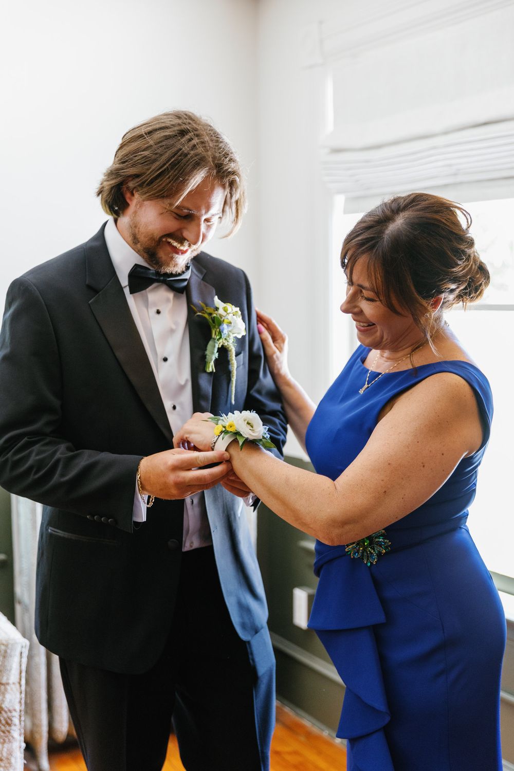 A groom in a black tuxedo smiles as a woman in a royal blue dress pins a boutonniere on his lapel.