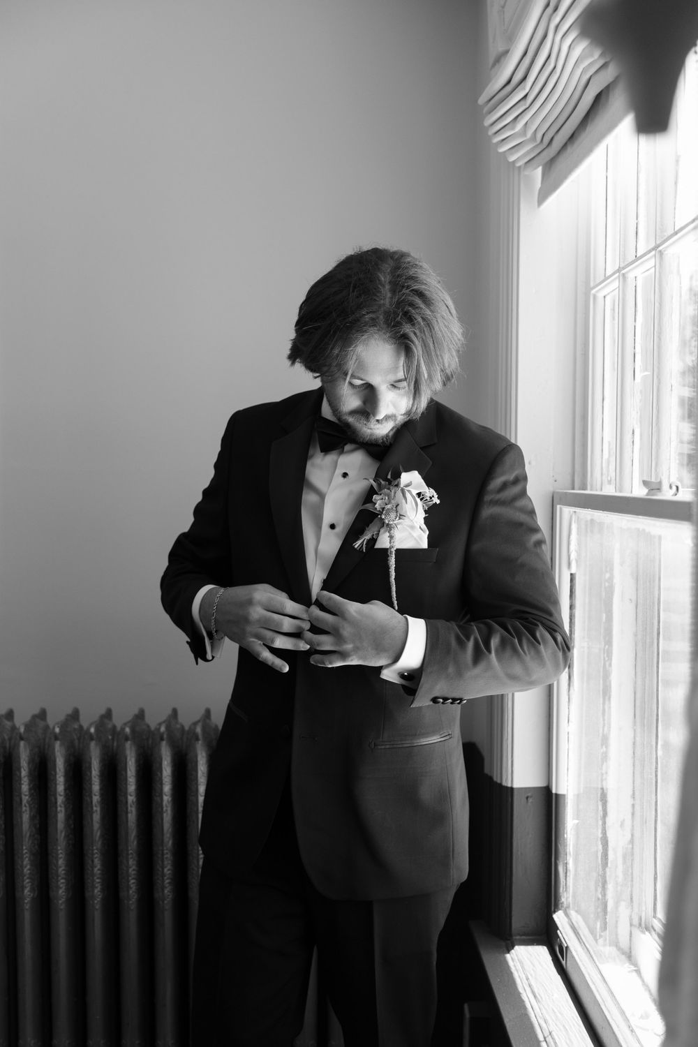 Groom in black tuxedo with boutonniere adjusting cufflinks by window in black and white wedding photo.