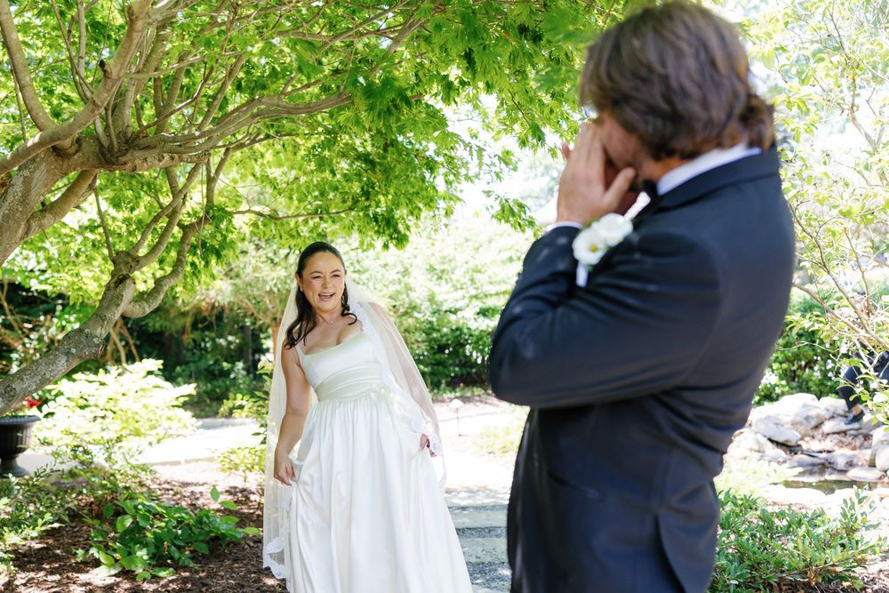 Bride in white gown smiles at emotional groom in garden during outdoor wedding first look ceremony.
