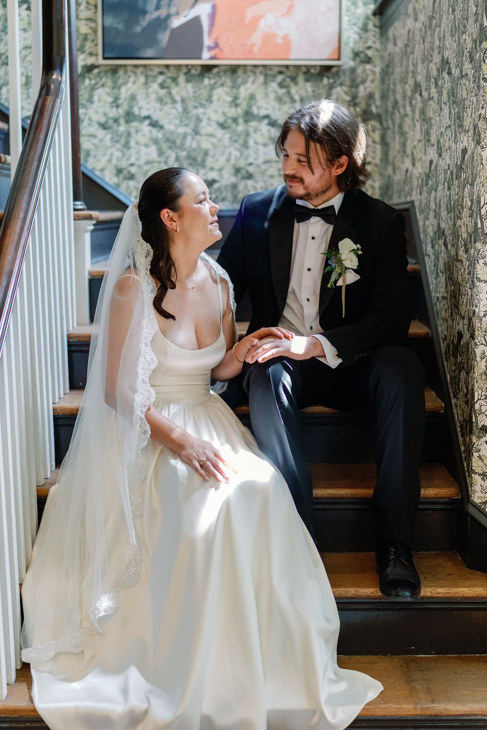 A bride in a white gown and veil sits with a tuxedo-clad groom on wooden stairs, sharing a loving glance.