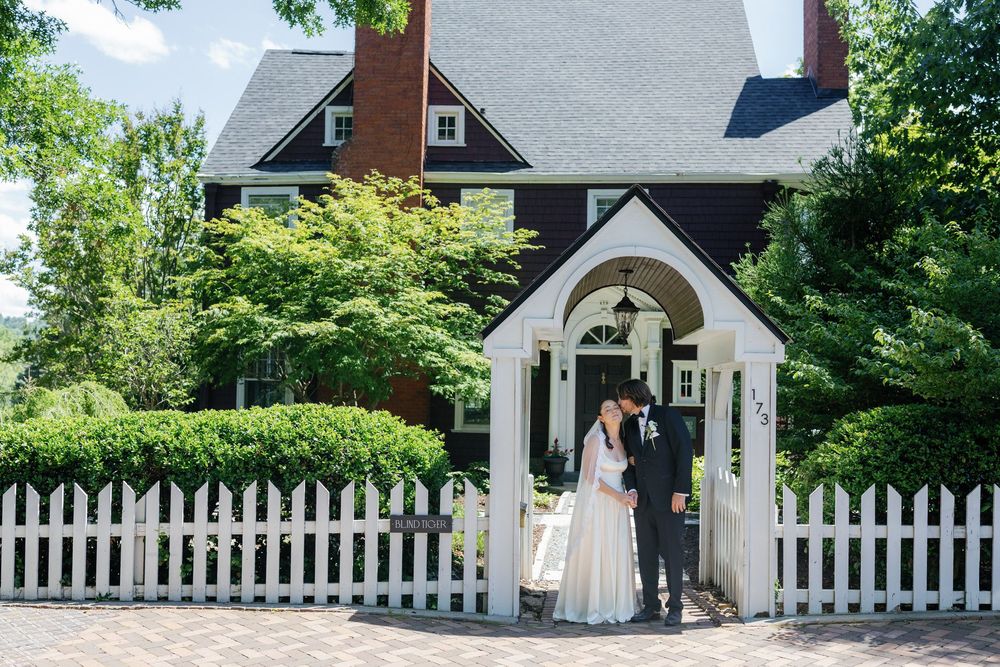 Bride and groom sharing a romantic kiss under a white arbor gate in front of a charming dark red colonial home.