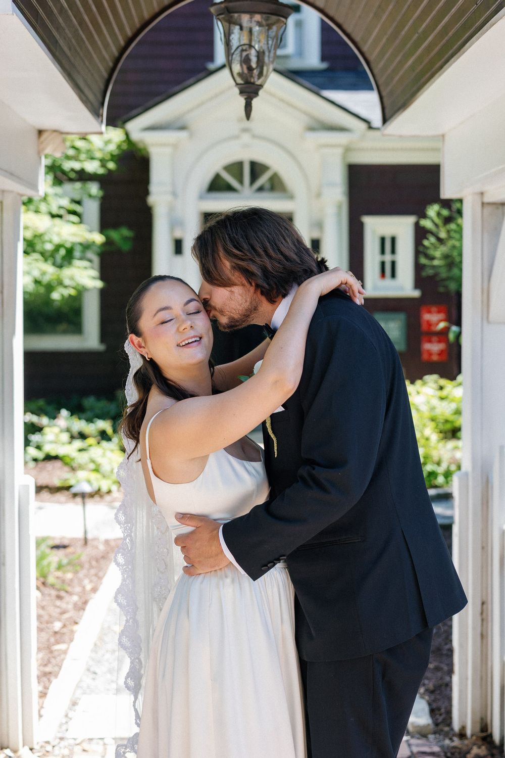 A bride in a white gown smiles joyfully as the groom in a black suit kisses her cheek outdoors.