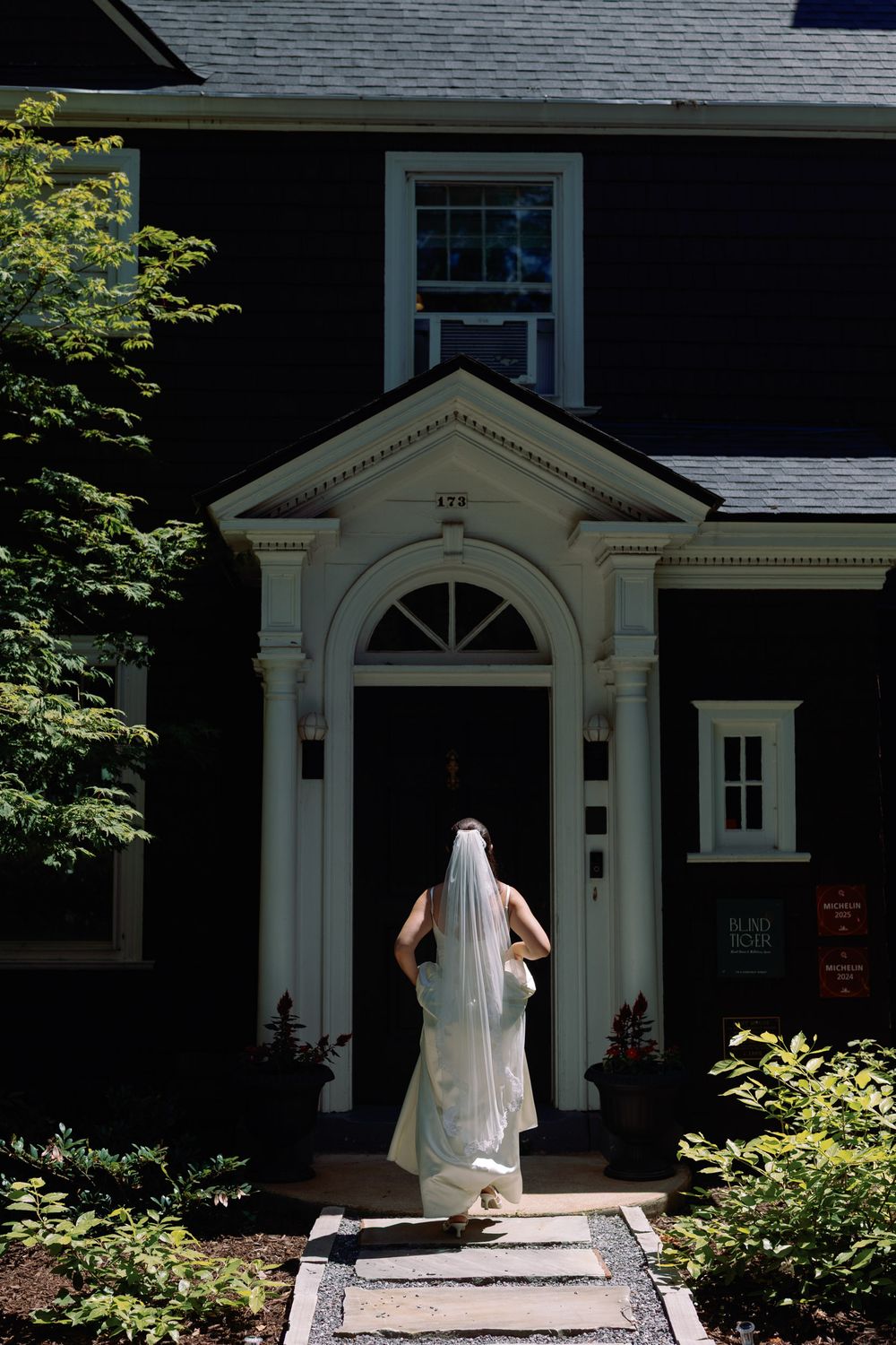 Bride in white gown and veil approaches elegant dark colonial home entrance on her wedding day.