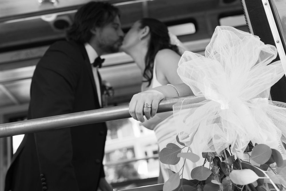 Black and white photo of a bride and groom kissing on a bus, with bridal bouquet in foreground.