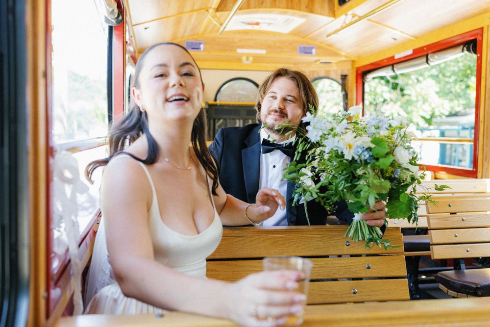 Happy bride and groom riding a vintage trolley, holding a white floral bouquet on their wedding day.