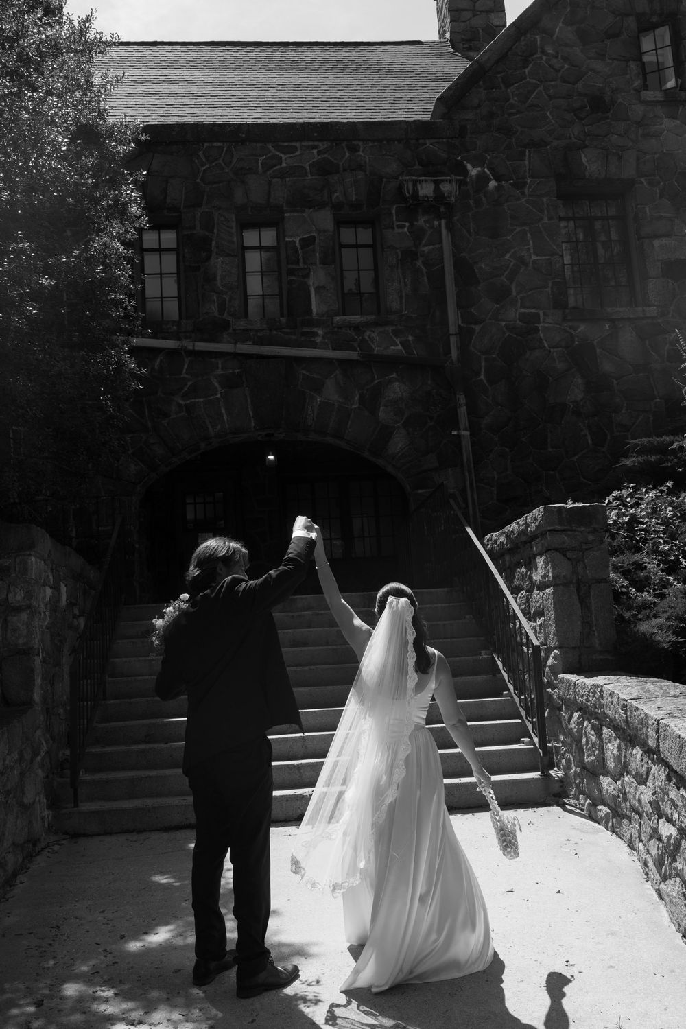 Black and white photo of bride and groom raising hands outside a stone castle venue on their wedding day.
