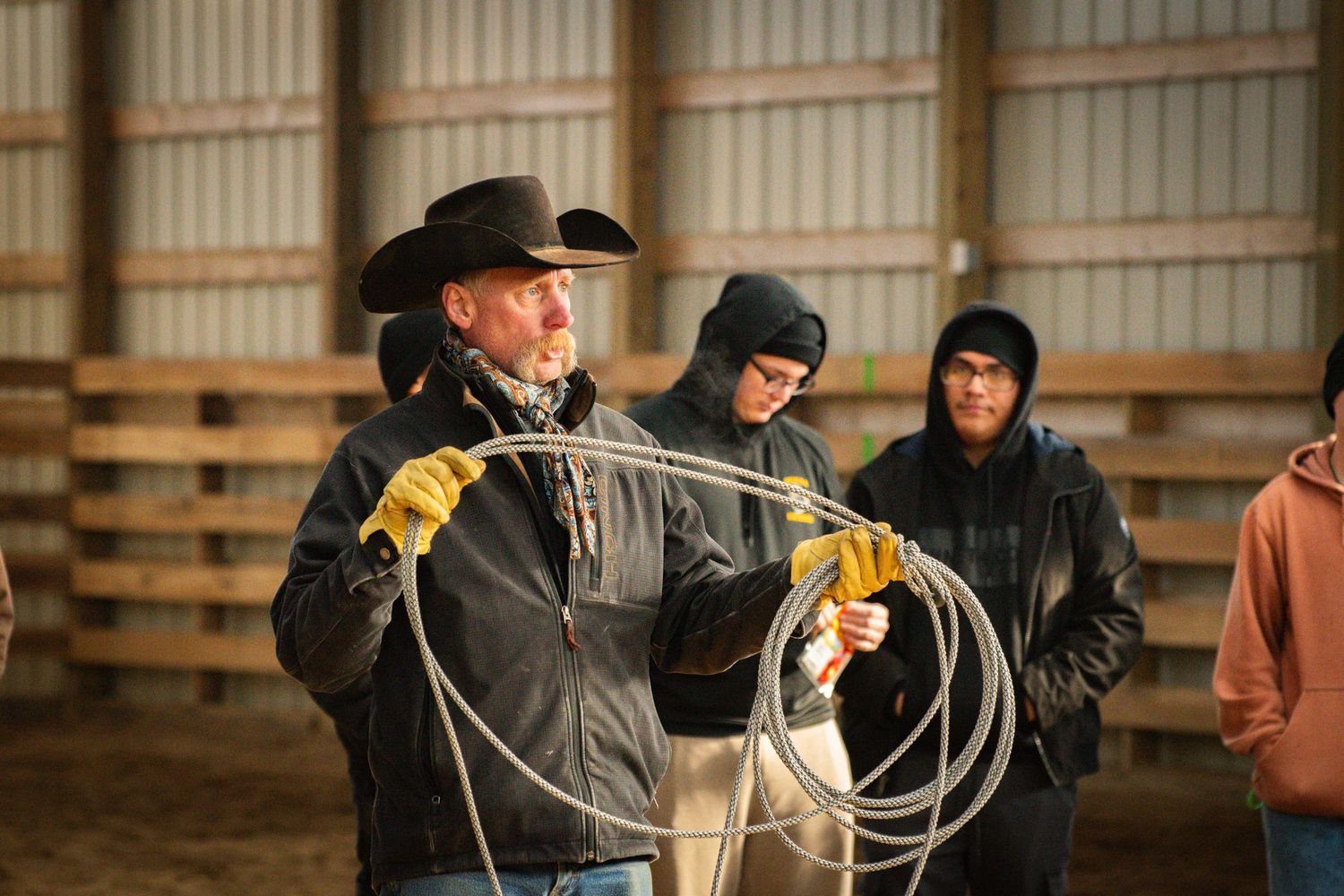 Cowboy instructor demonstrating rope handling techniques to students inside a barn.