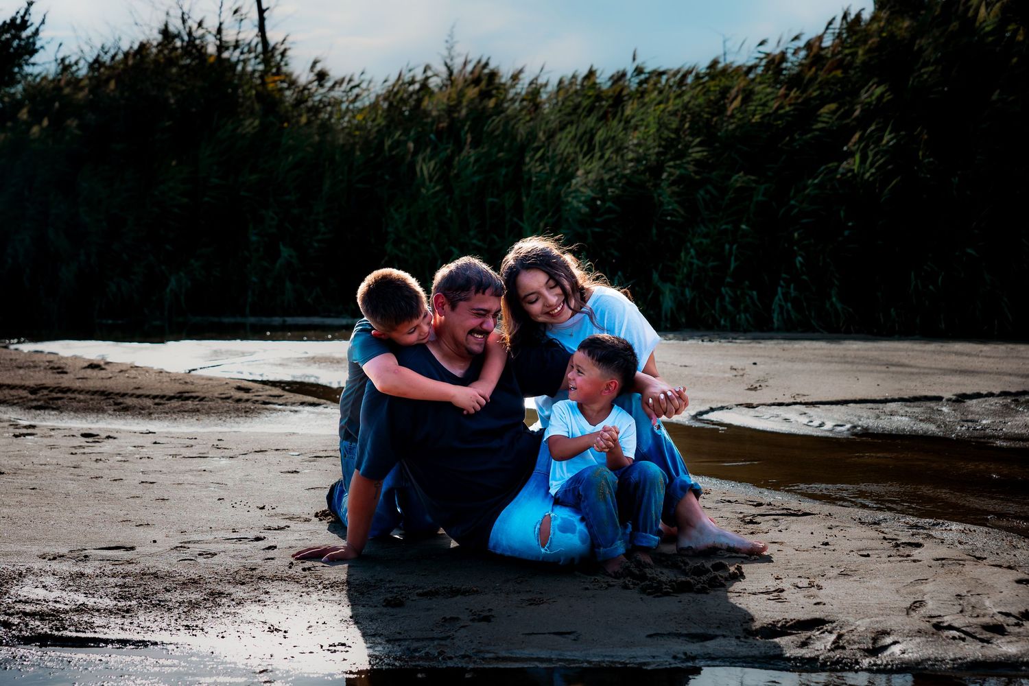 Family of four laughing and cuddling together on a sandy beach with lush green trees in the background.