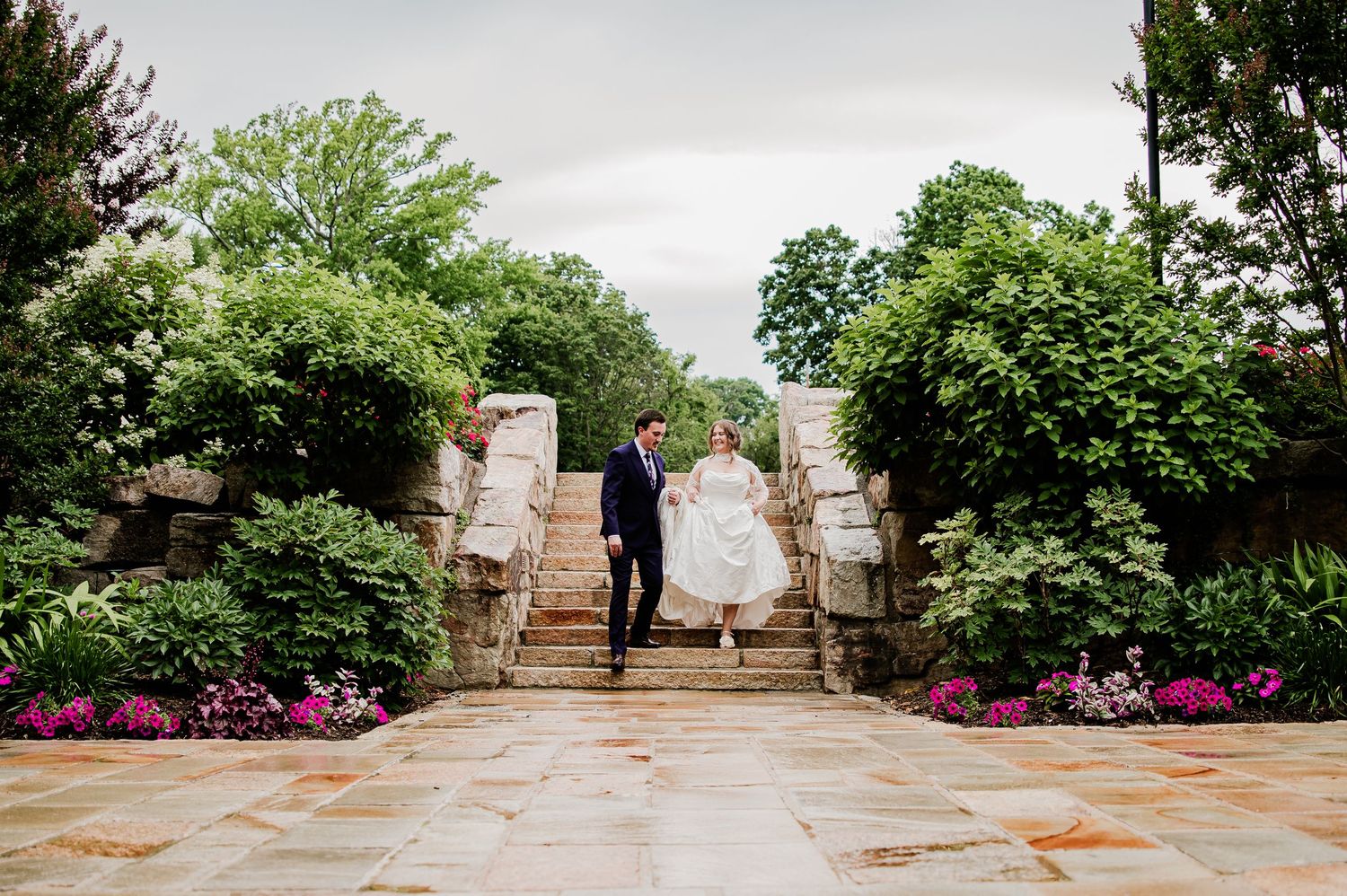 Bride and groom walk down stone garden stairs surrounded by lush greenery and pink flowers at an outdoor wedding venue.