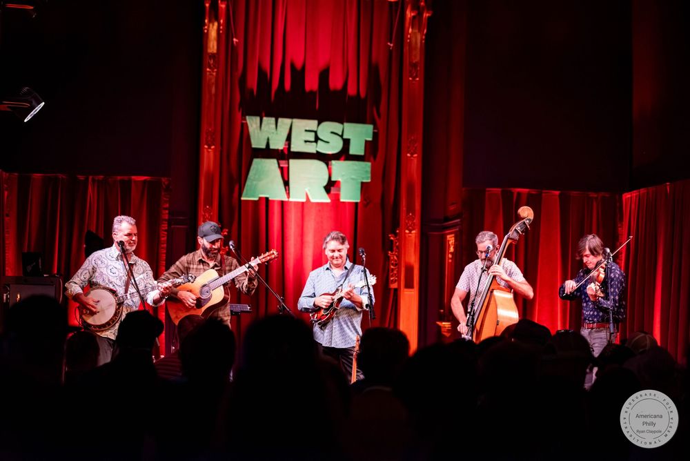 Five musicians perform bluegrass on stage at West Art venue, playing banjo, guitar, and upright bass under red stage lighting.