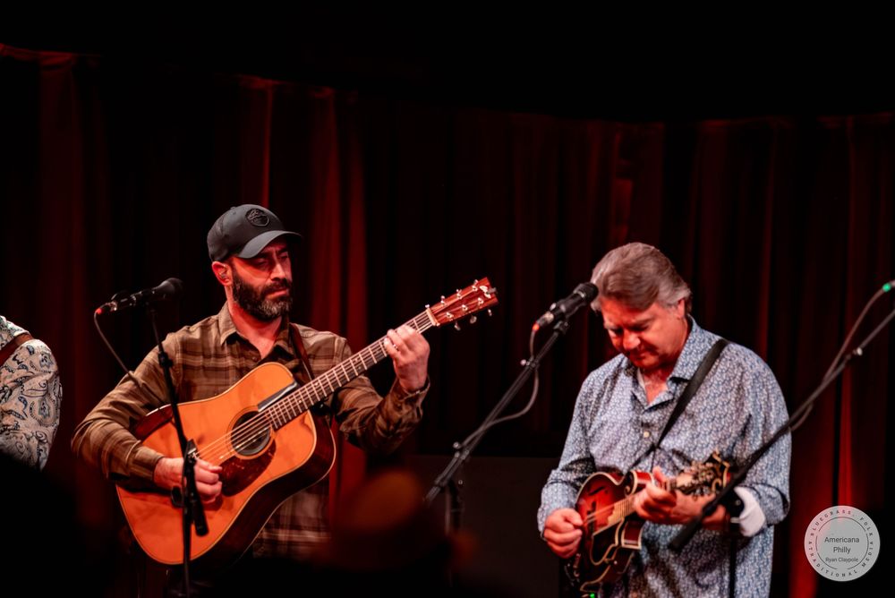 Two musicians perform on stage, one playing acoustic guitar and another on mandolin, at a live bluegrass concert.