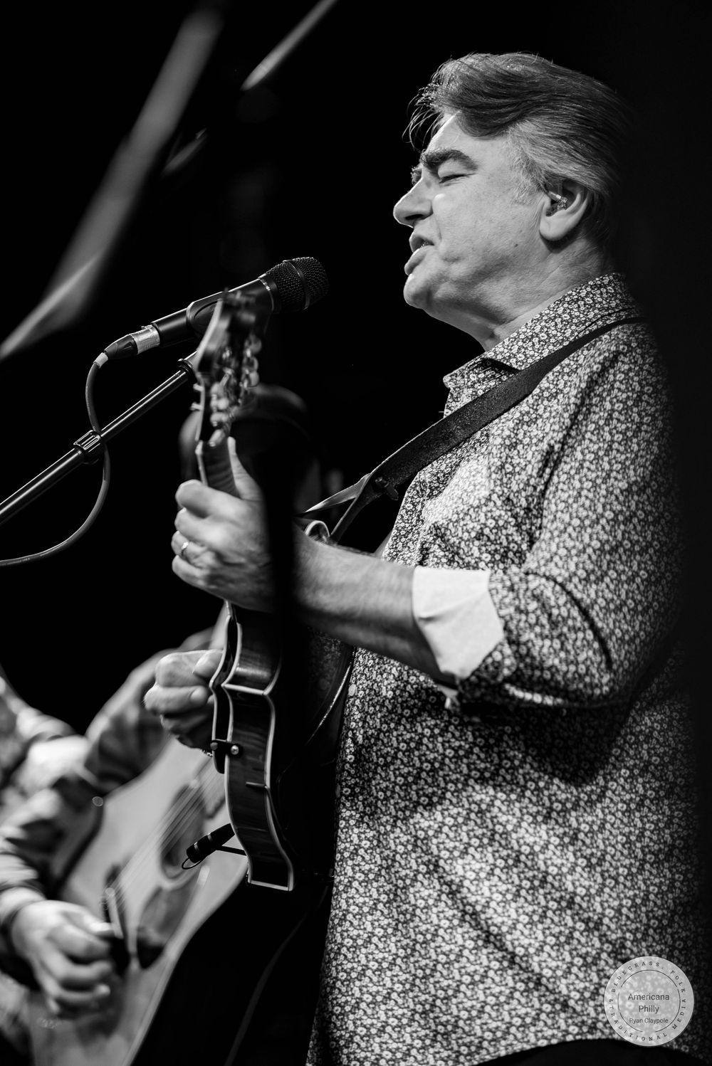 Black and white photo of a man playing mandolin and singing into a microphone on stage.
