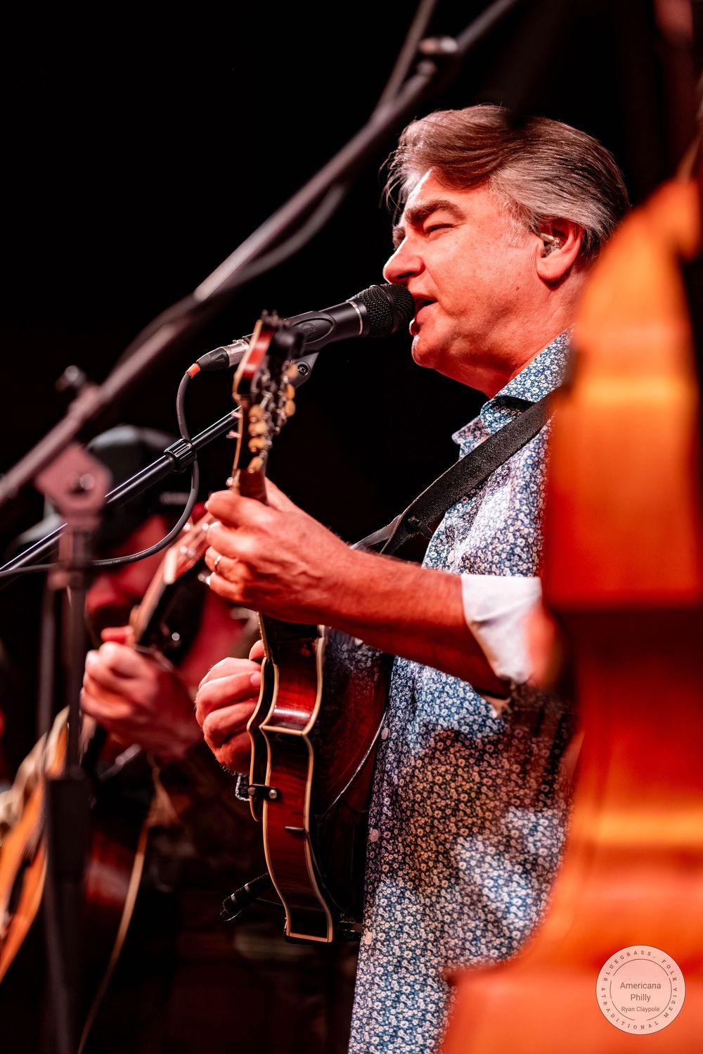 Man playing mandolin and singing into microphone on stage wearing floral shirt at live music performance.
