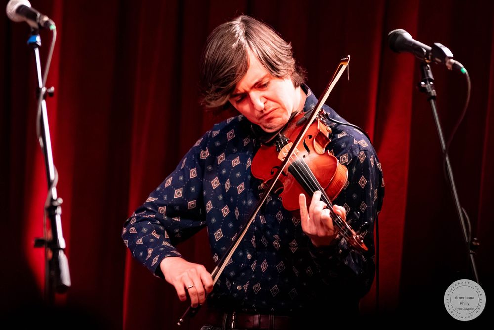 Male violinist in floral shirt performs on stage with red curtain backdrop and microphone stands nearby.