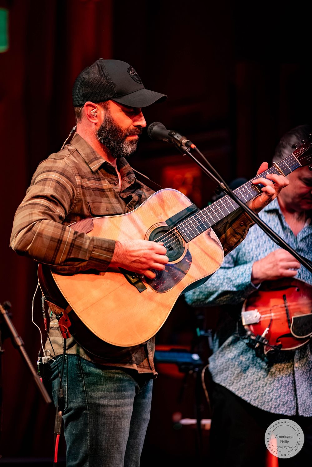 Bearded man in plaid shirt and black cap playing acoustic guitar on stage with microphone at live music event.