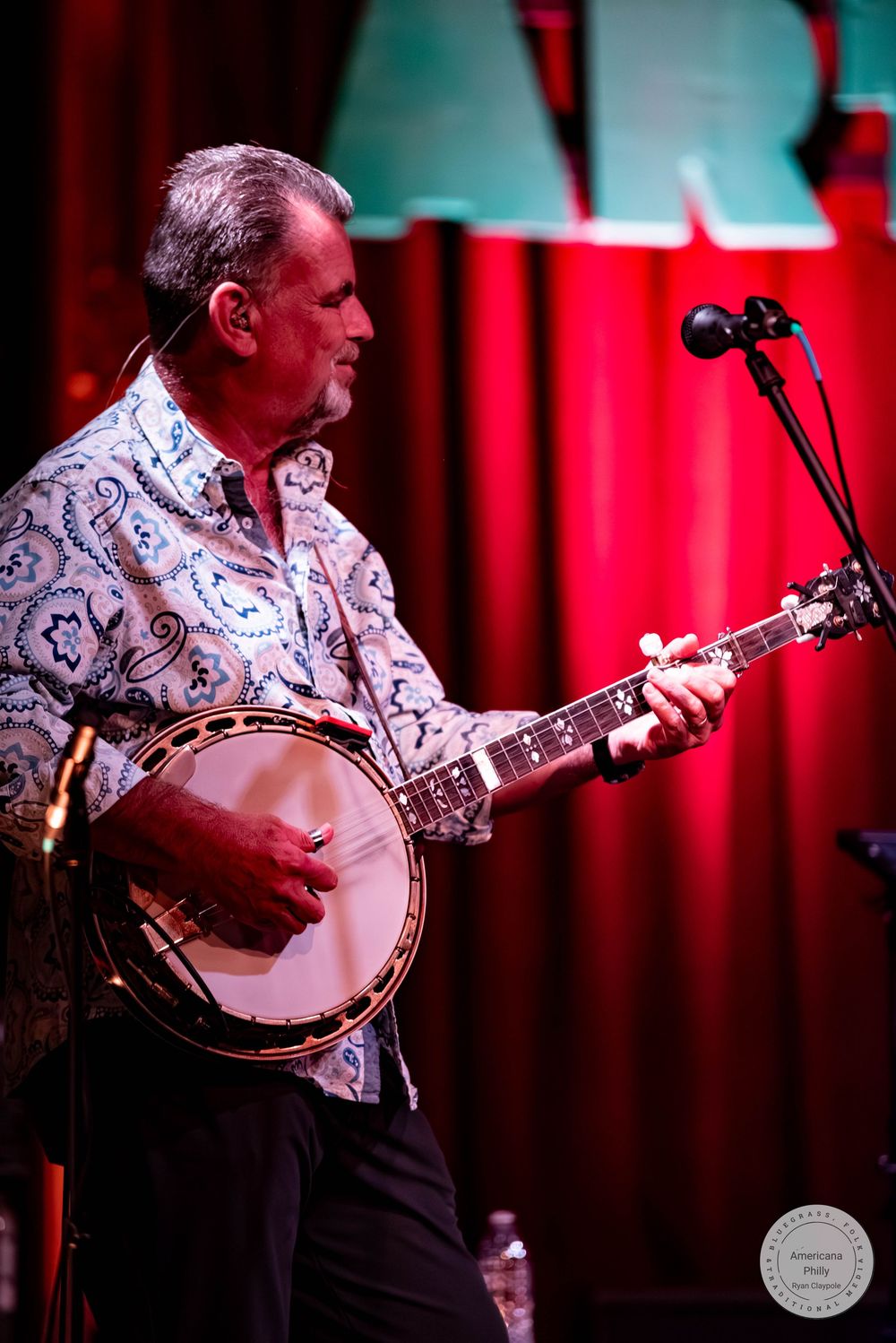 A man plays a banjo on stage, wearing a floral shirt under red stage lighting with a microphone stand nearby.