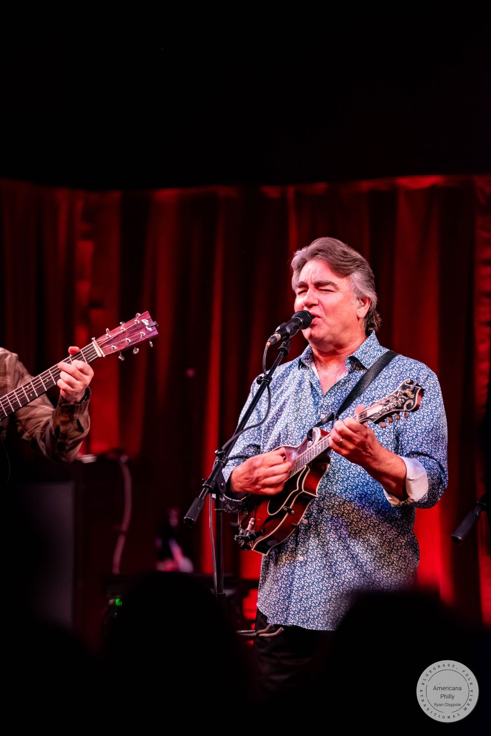 Man playing mandolin and singing into microphone on stage with red curtain backdrop at live music performance.