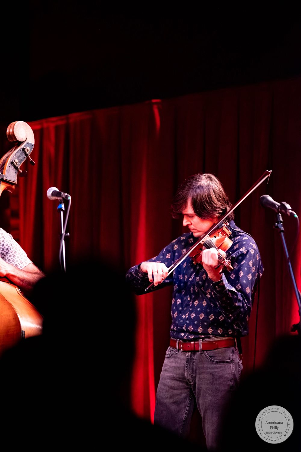 A musician plays violin on a dimly lit stage with red curtains, wearing a blue patterned shirt and jeans.