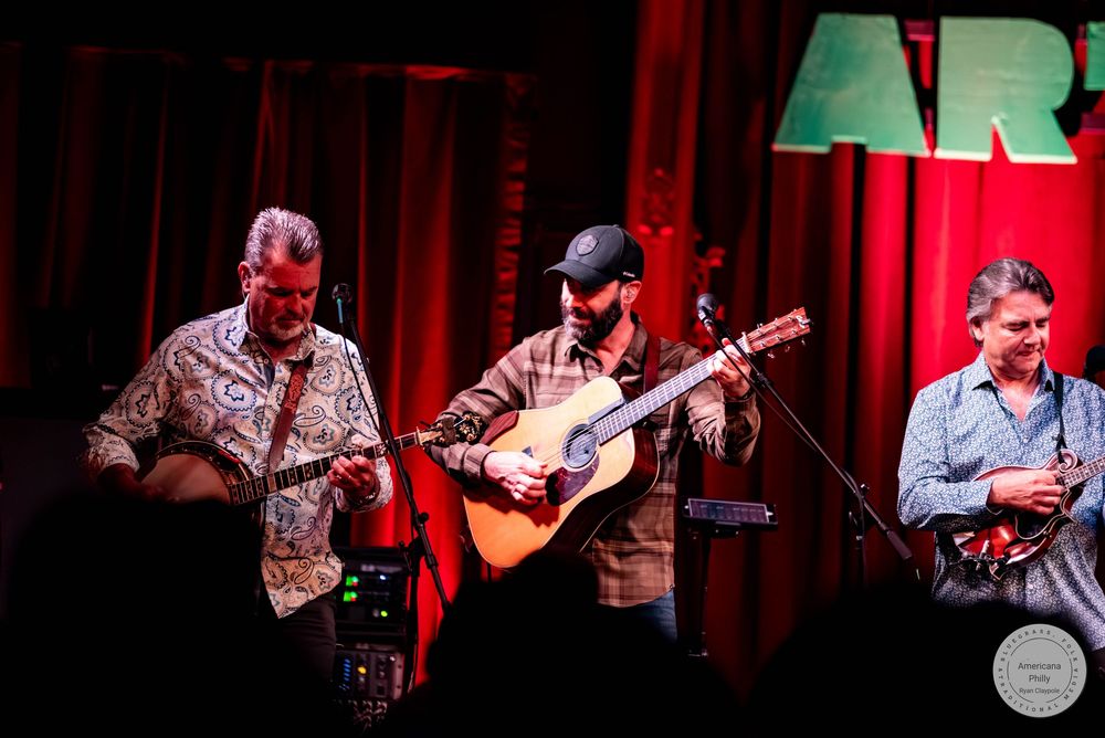 Three musicians perform live on stage, two playing guitars and one with a mandolin, under red stage lighting.