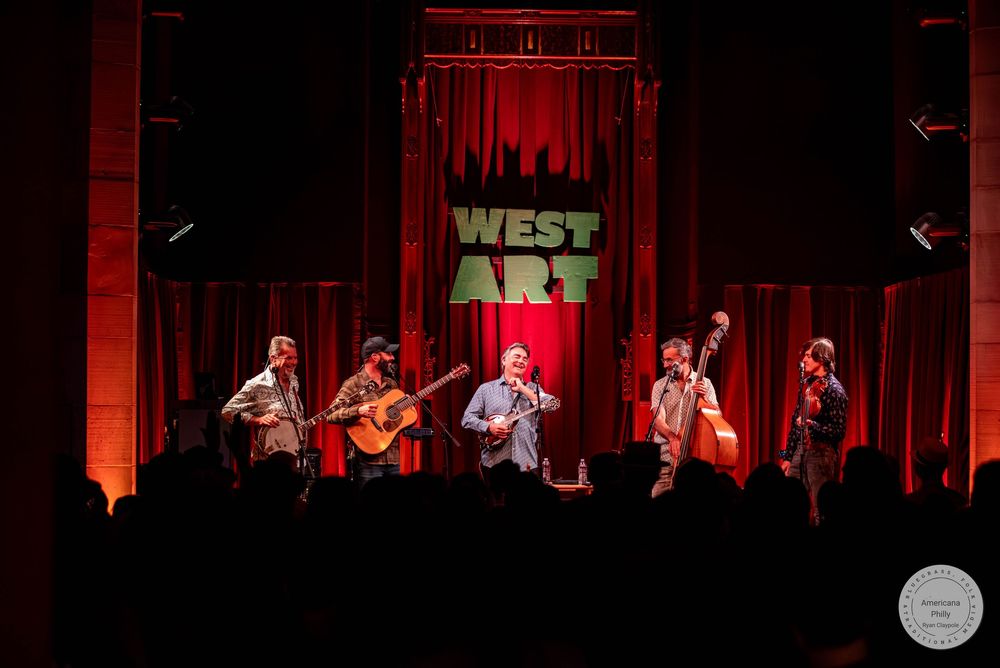 Five musicians performing live on stage at West Art venue, bathed in warm red lighting.