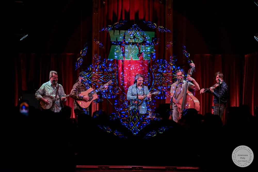 Four musicians perform bluegrass on stage with colorful stained glass backdrop and red curtains at West End venue.