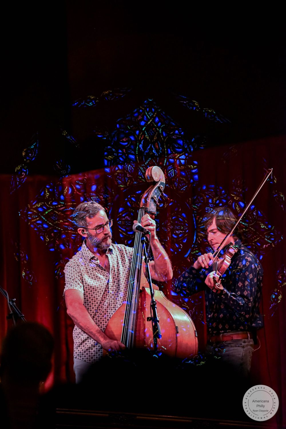 Two musicians perform live, one playing upright bass and another playing violin on a dimly lit stage.