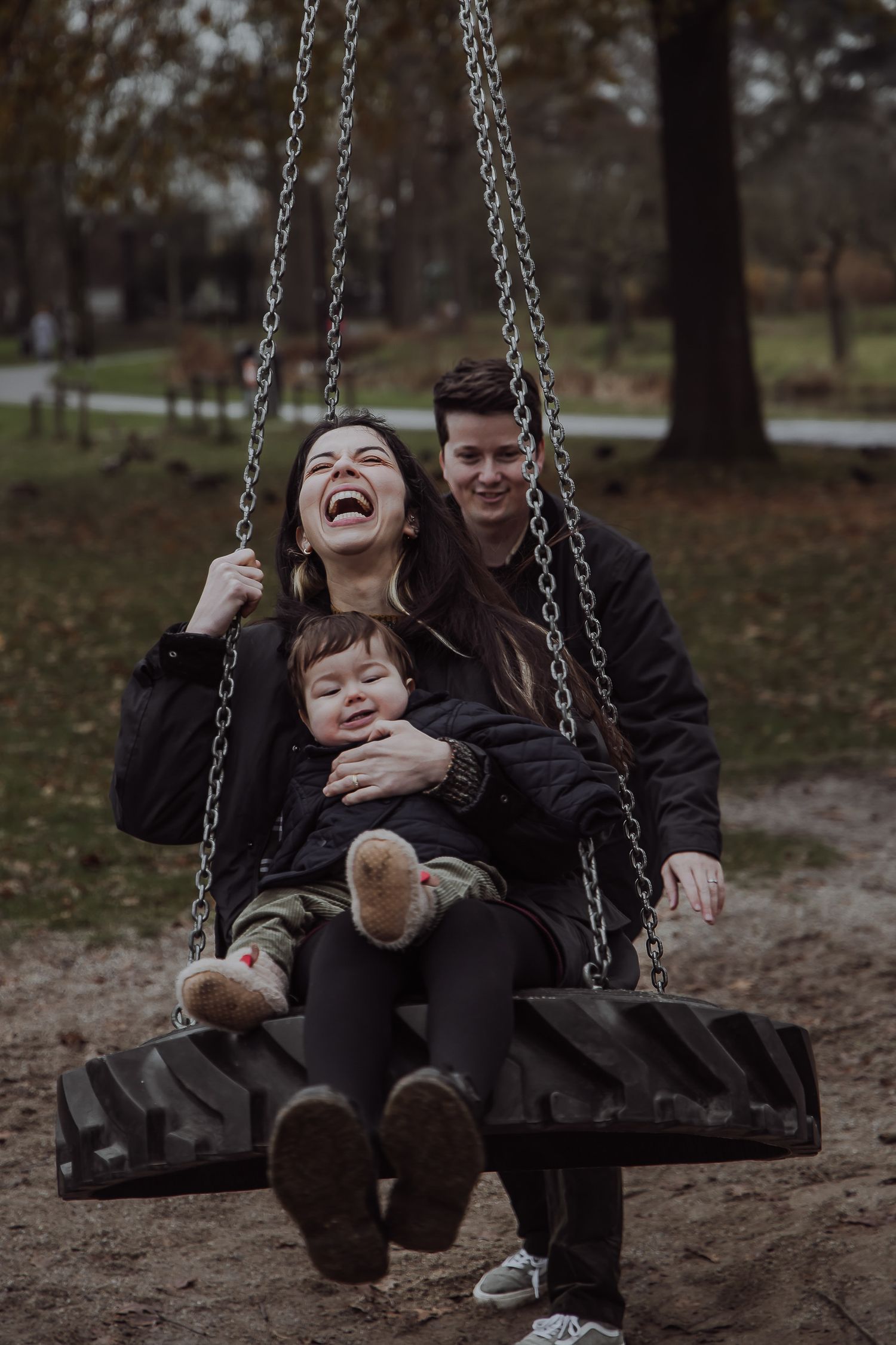 Family laughing together as dad pushes mum and baby on a swing during an outdoor winter photoshoot in Woerden