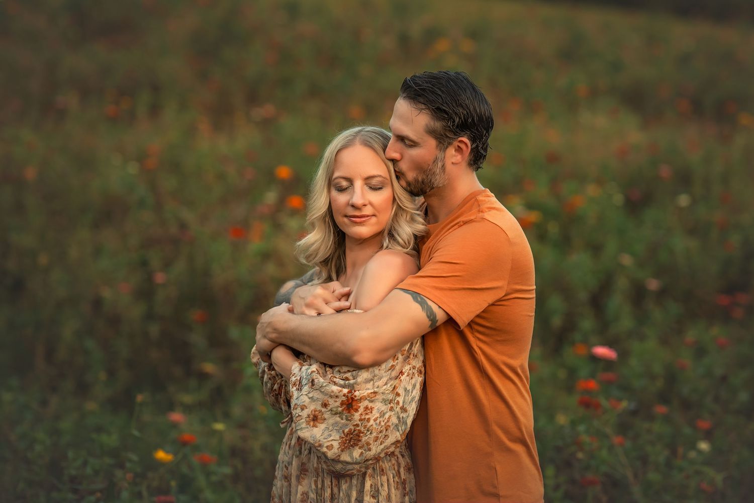 Couple embracing in a flower field at golden hour, man in orange shirt kissing womans head tenderly.