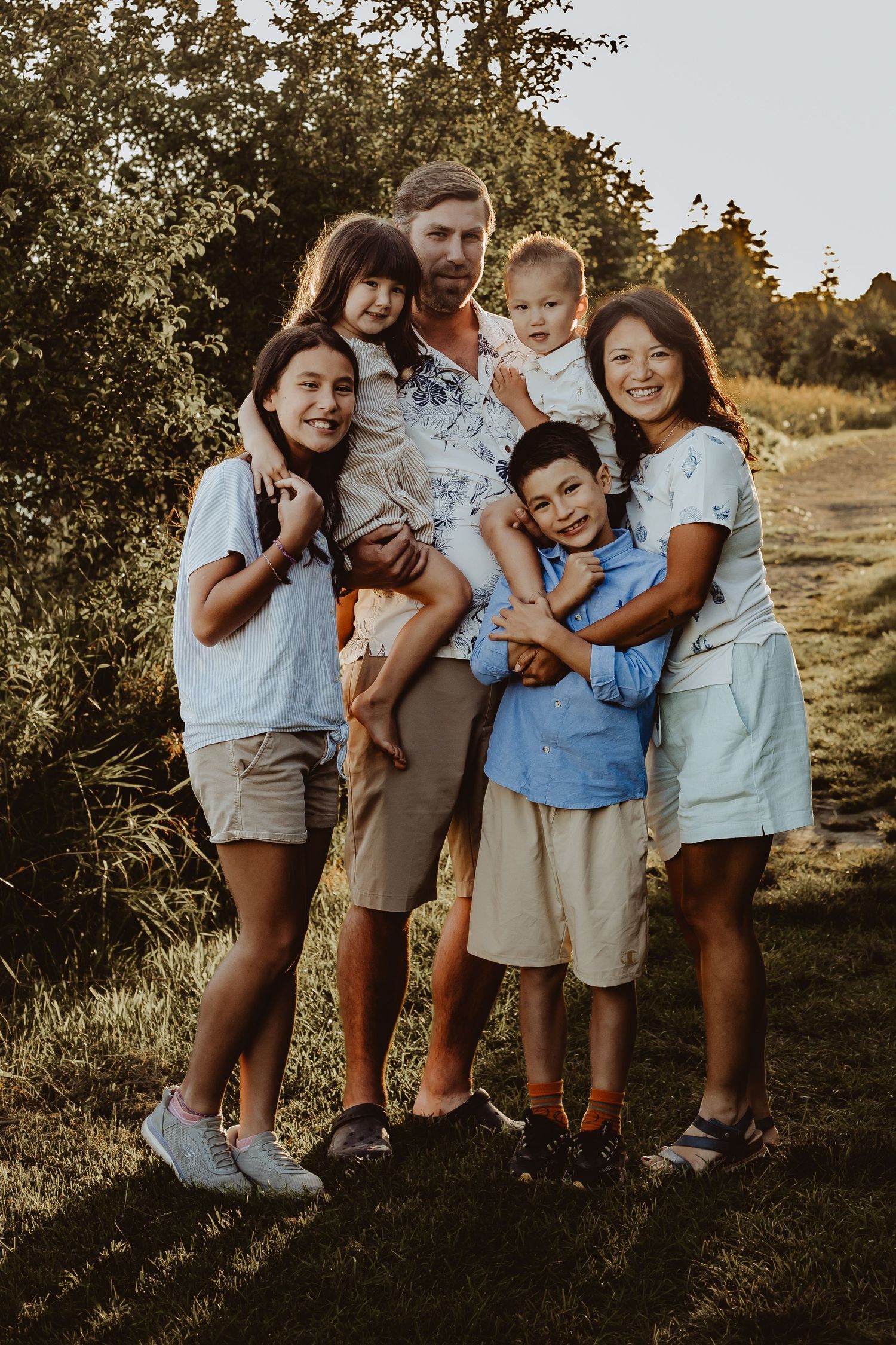 Happy family of six posing outdoors at golden hour, parents with four children smiling in a lush green park setting.