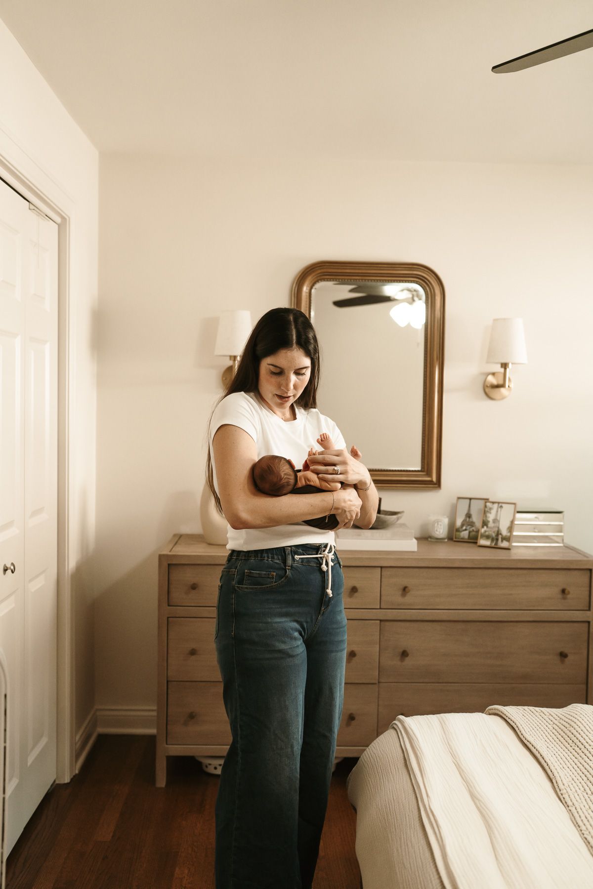 Mom in jeans holding a baby in a minimalist bedroom with wooden furniture in Mount Prospect
